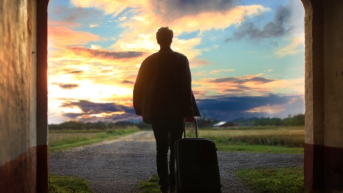 A man standing in a concrete tunnel with a suitcase looking out as the sun is setting.