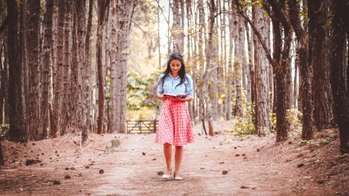 A young woman reading a book while walking on path lined by trees.