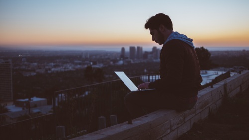 A young man sitting on the edge of wall with a laptop on his lap.
