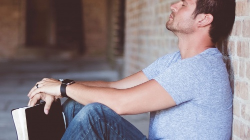 A man holding his Bible with his back propped against a wall - with eyes close and head down praying.