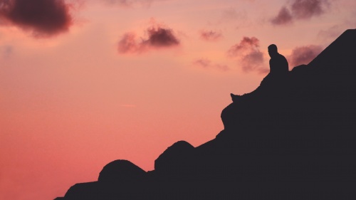 A person sitting a hill with a red sky.