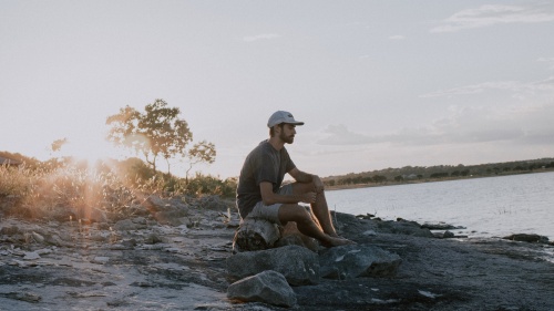 A man sitting on a log by a lake. 