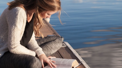 A woman sitting on a dock in the water reading her Bible.