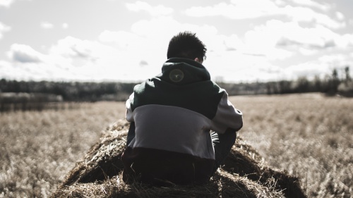 A young man sitting on a round bale of hale looking out over a field. 