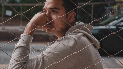 A young man sitting next to chain fence with his head on his hand.