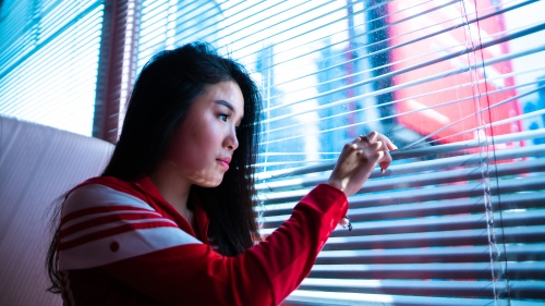 A young woman looking through the blinds on a window.