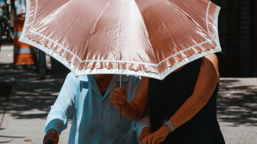 Photo of two women walking under a parasol; an elderly woman with a walker being helped by a middle-aged woman.