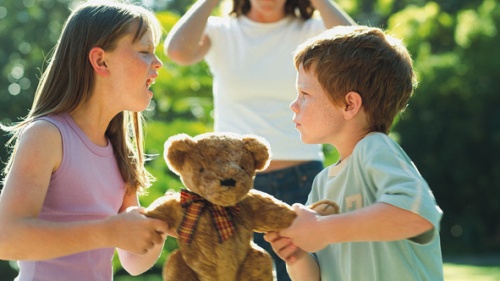 Two siblings fight over a stuffed teddy bear with the mom in the background.