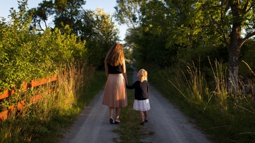 Photo of a mother and young daughter walking down a dirt road in the countryside; the daughter is holding her mother's hand and looking up at her.