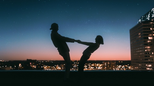 a man and woman in silhouette against a city skyline and starry sky, holding hands and leaning back mid-twirl