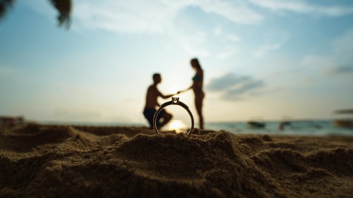 silhouette of man proposing to woman on a beach seen through a ring up close