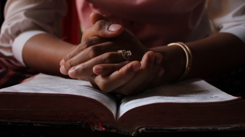 a pair of hands folded in prayer sitting atop an open Bible