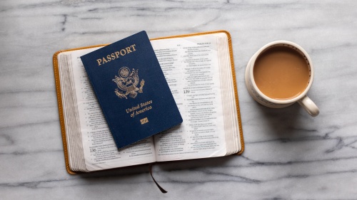 a open Bible with a US passport placed on top sitting beside a mug of coffee on a marble tabletop