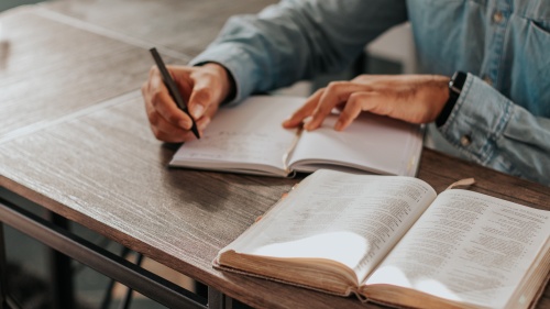 A man writing in a notebook with a Bible open.