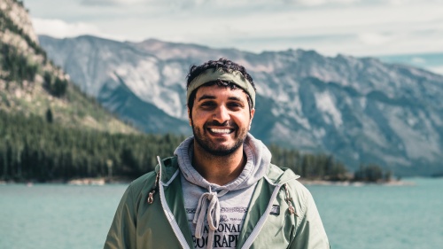 a man stands outdoors with mountains and a lake behind him