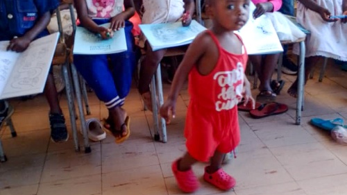 a group of children seated indoors, with one small child walking in front of them