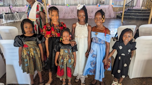 a group of girls wearing fancy dresses standing indoors with a lady walking the background