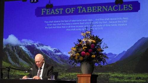 a man standing at the podium with a Feast of Tabernacles powerpoint presentation projected behind him