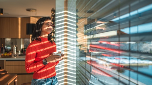 a woman standing indoors and looking out through a reflective window while holding a coffee cup