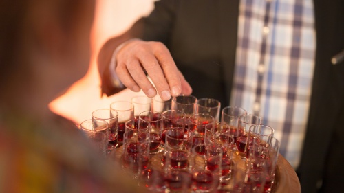 a man's hand reaching for a shot glass with red wine on a tray of similar glasses