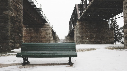 A park bench surrounded by snow and underneath a bridge.