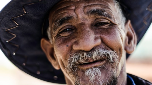 Upclose photo of a older man with smile and grey goatee - wearing a hat.