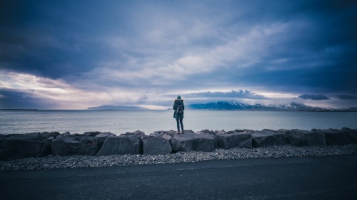 W woman standing on big rocks looking out at cold water.