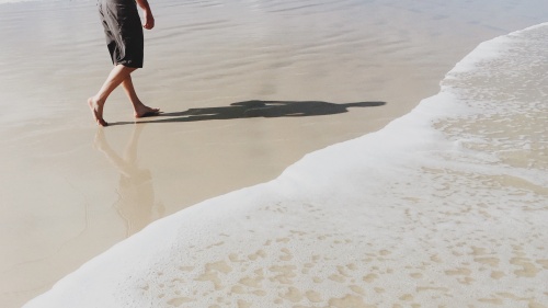 A man walking on the beach beside the ocean wave. His shadow on the flat sand.