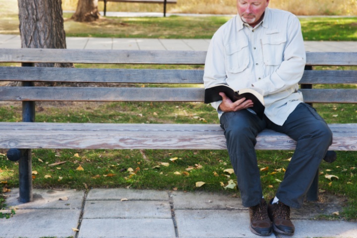 A man reading a Bible sitting on a park bench.