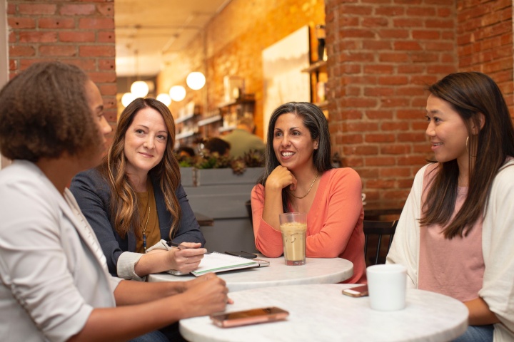 a group of four women dressed in business casual sitting at a table in a cafe with a brick wall that has a window looking into the rest of the cafe