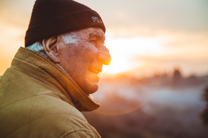 an elderly man wearing a hat and coat as he stands outdoors looking into the distance with a sunset in the background