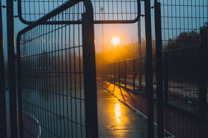 A metal fence with its gate opening. The sun is rising in the background.