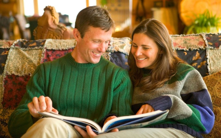 A couple sitting together on a couch looking at a large book.