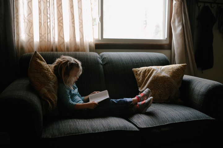 A little girls sitting on a couch looking at a book.