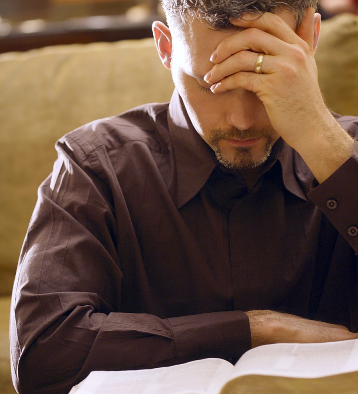 A man with his hand holding his head while looking down at a Bible.