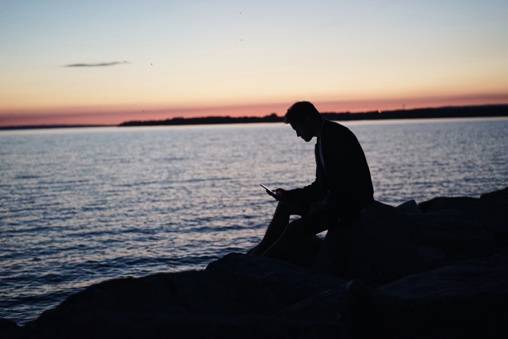 A man sitting on rocks by a body of water looking down at his phone.