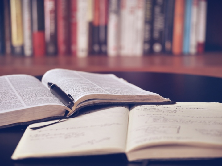 A open Bible laying on a table with pen inside of it and notebook beside it.