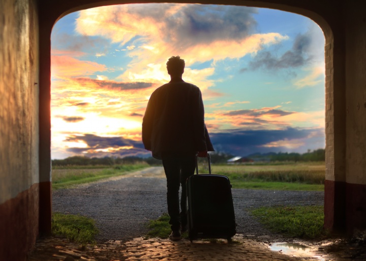 A man standing in a concrete tunnel with a suitcase looking out as the sun is setting.