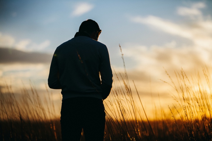 A man looking down while standing in a field. The sun is setting in the distance.