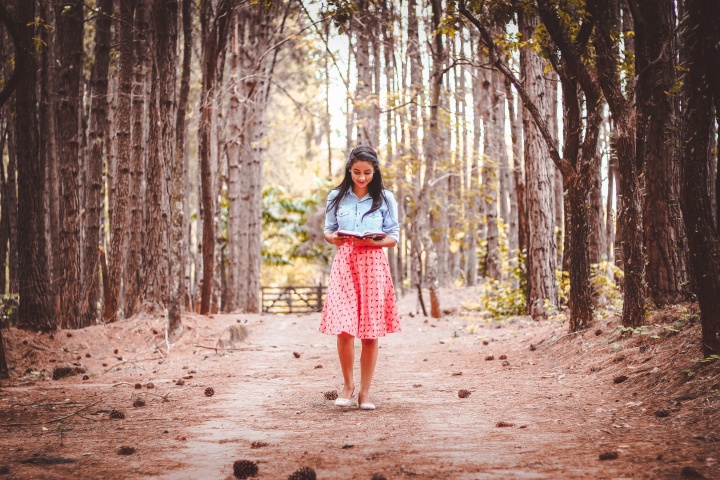 A young woman reading a book while walking on path lined by trees.