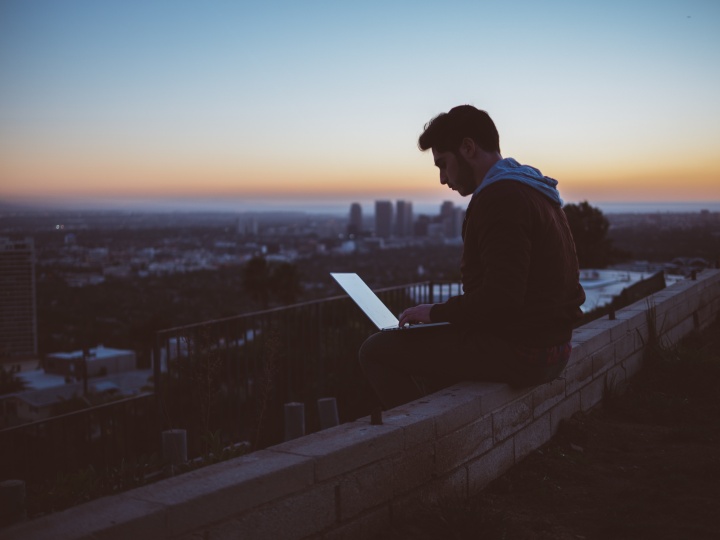 A young man sitting on the edge of wall with a laptop on his lap.