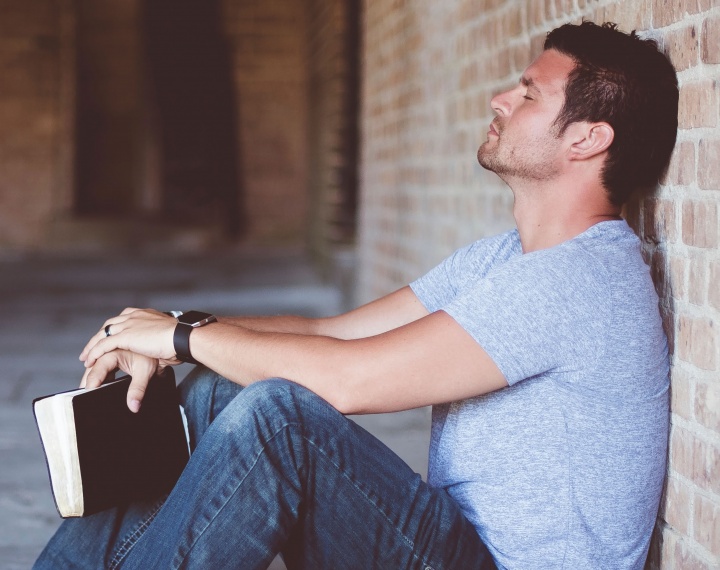 A man holding his Bible with his back propped against a wall - with eyes close and head down praying.