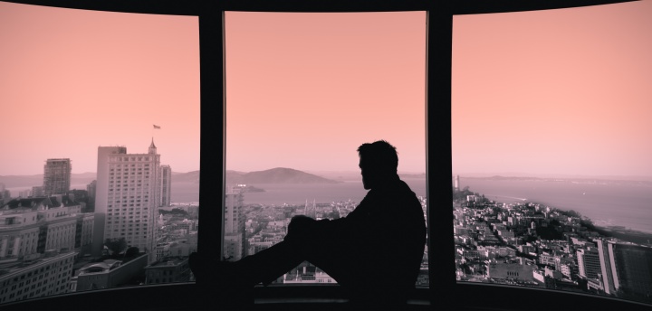 A man sitting in a window sill looking out over a city skape.