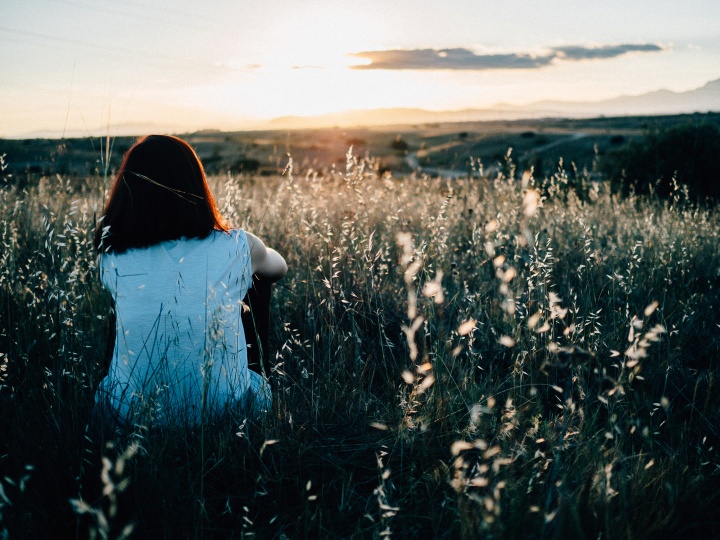 A young woman sitting in a field of grass and flowers watching the sunset.