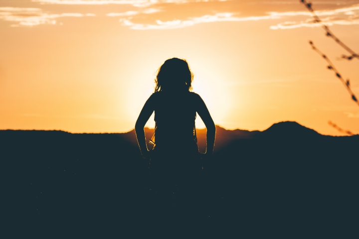 A silhouette of young woman with small mountains and a setting sun in background.