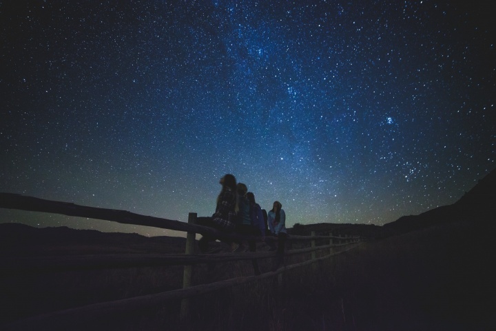 People sitting on a fence looking up at the night sky full of stars.