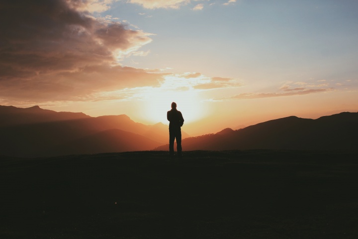 A silhouette of a person stand on a hill with the sun in the background.