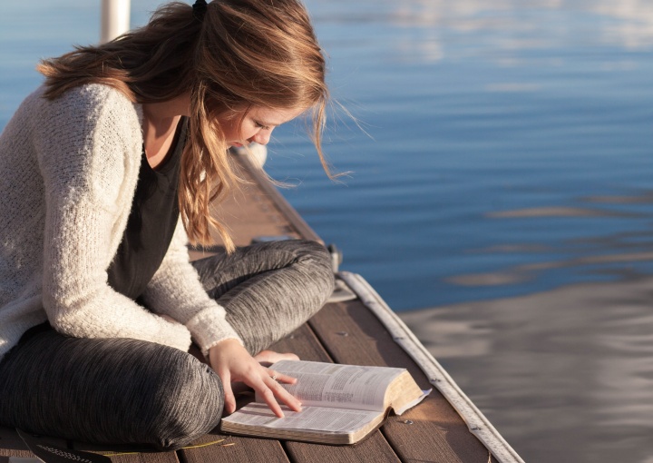 A young woman reading a Bible on a dock in the water.