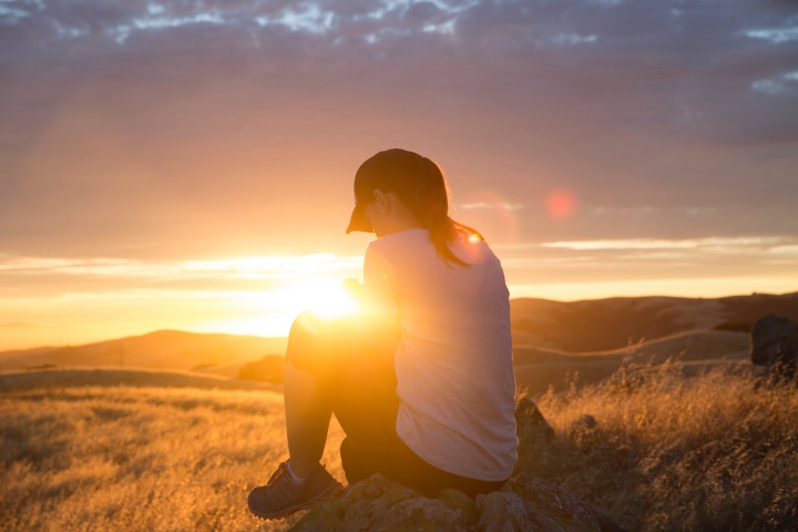 A woman praying while sitting on a rock with the sun setting in the background.