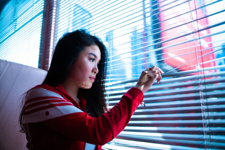 A young woman looking through the blinds on a window.